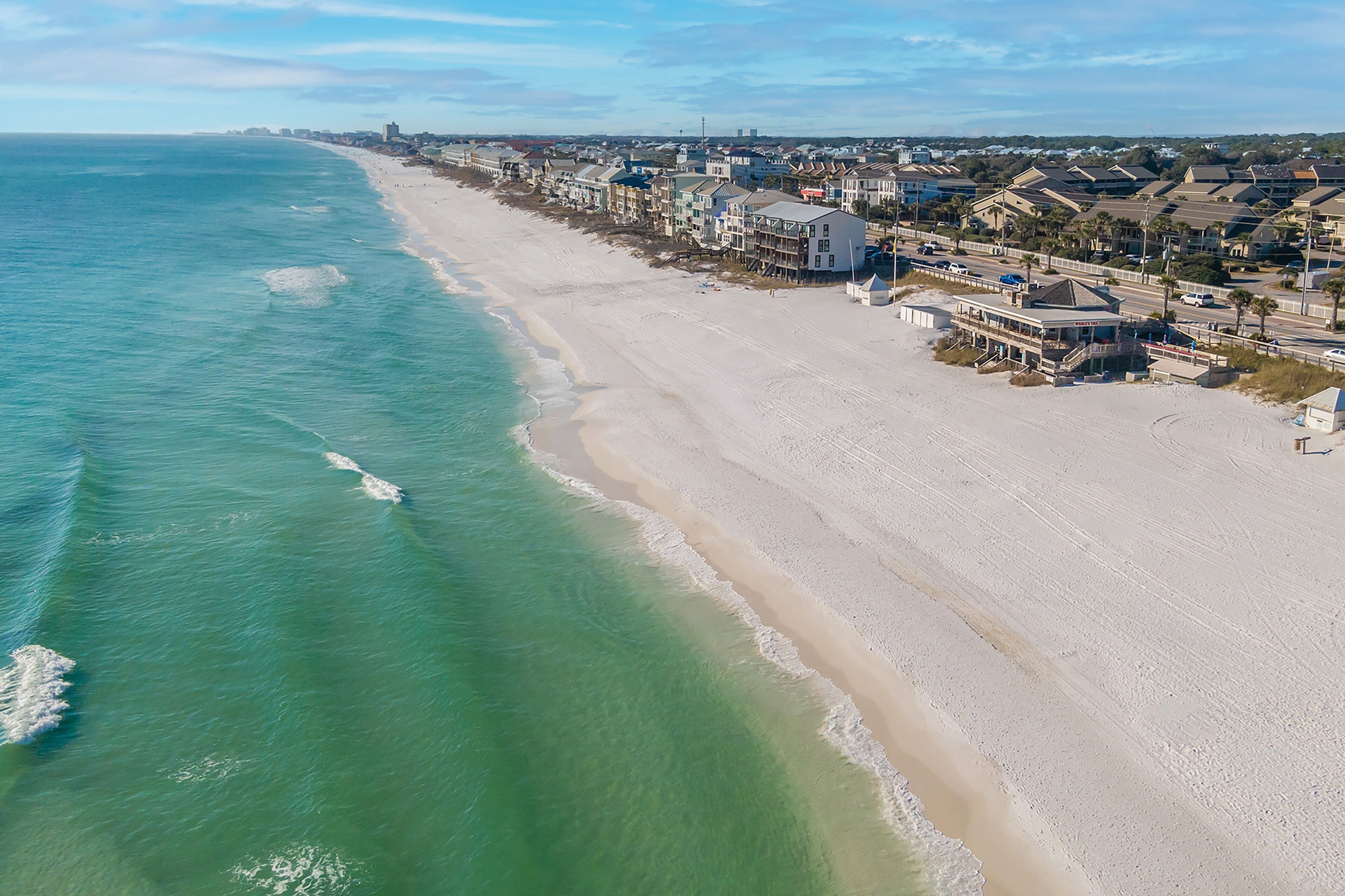 An aerial view of a coastal area with a beach, beachfront properties, and a road running parallel to the shore.