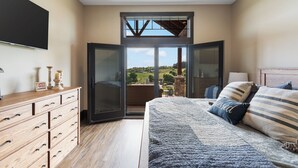 A bedroom with a large bed, a wooden dresser, a TV, and a balcony with a view of the countryside. - The Cottage at Heritage Hill (Sugarcreek)