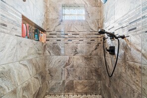 A shower with a handheld showerhead, a glass window, and a shelf with toiletries.