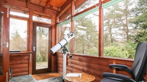 A wooden cabin with a telescope on a table, a chair, and a view of trees through large windows. - Rocksite (Blairgowrie)