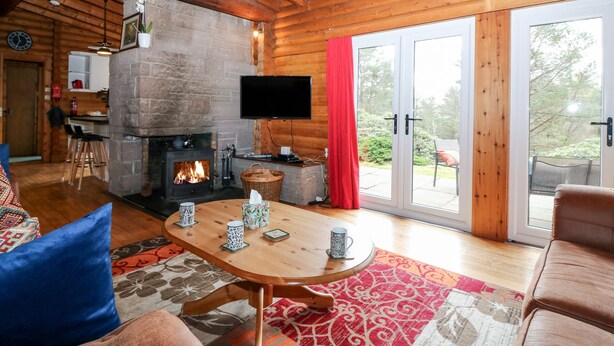 A cozy log cabin interior with a fireplace, a dining area, and a large sliding glass door leading to an outdoor patio. - Rocksite (Blairgowrie)