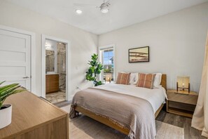 A bedroom with a large bed, a bedside table with a lamp, a plant, and a view of the outside. - Aurora 7 BR Elevator Theater Rec Room, Pool & Hot Tub (Corolla)