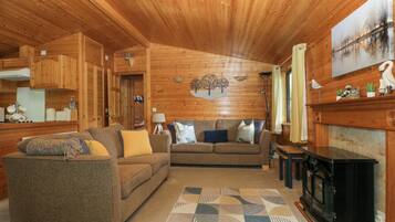 Een houten chalet van binnen met een bank, een kamerkleed met dessin, een open haard en een eethoek.