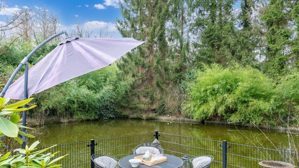 Patio esterno con tavolo e sedie, un ombrellone viola e vista su un fiume, alberi e una recinzione.