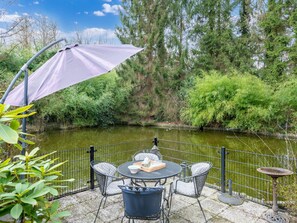 An outdoor patio with a table and chairs, a purple umbrella, and a view of a river with trees and a fence. - Arendonk Pondside Chalet (Arendonk)