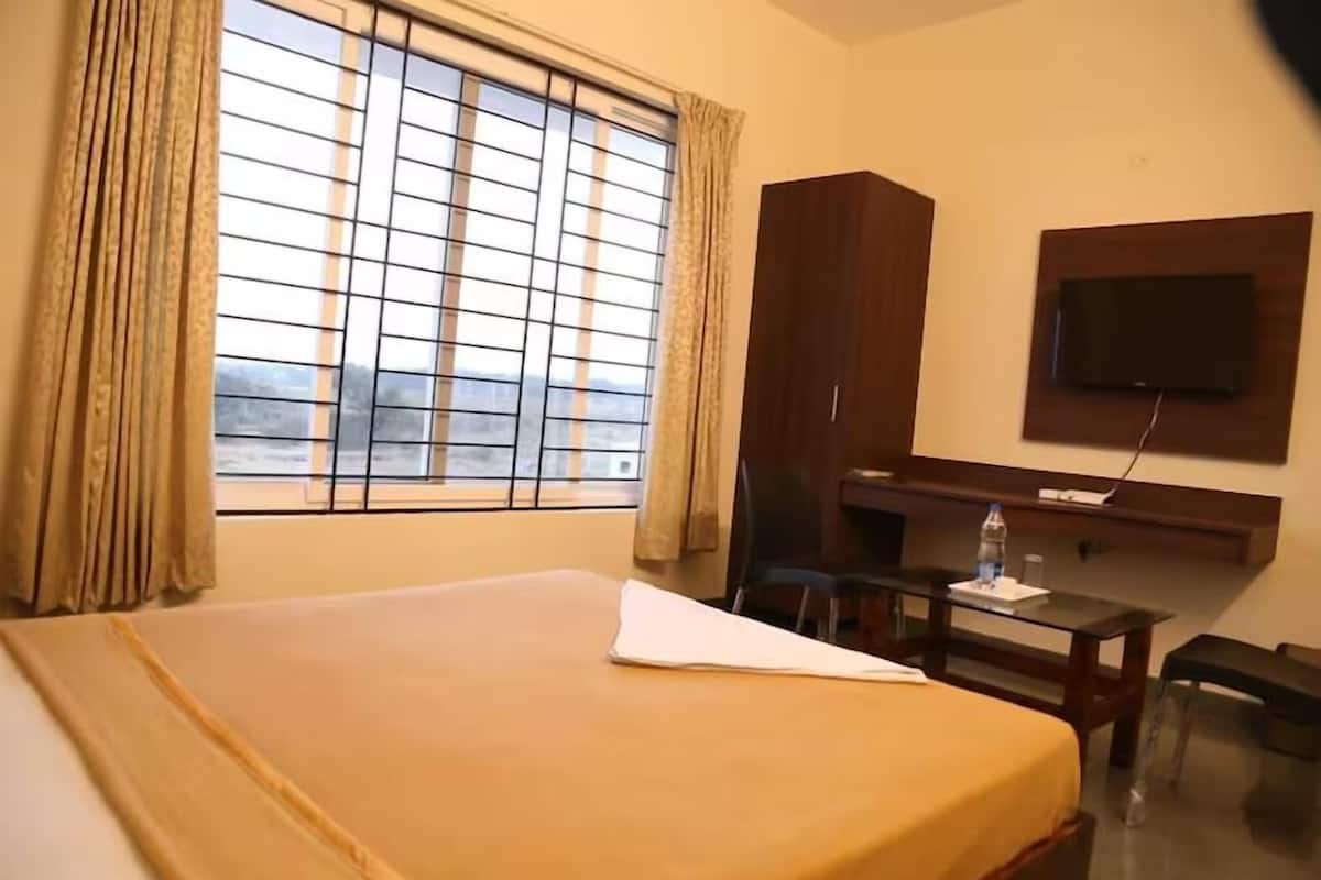 A hotel room with a bed, a desk, a chair, a TV mounted on the wall, and a window with curtains.