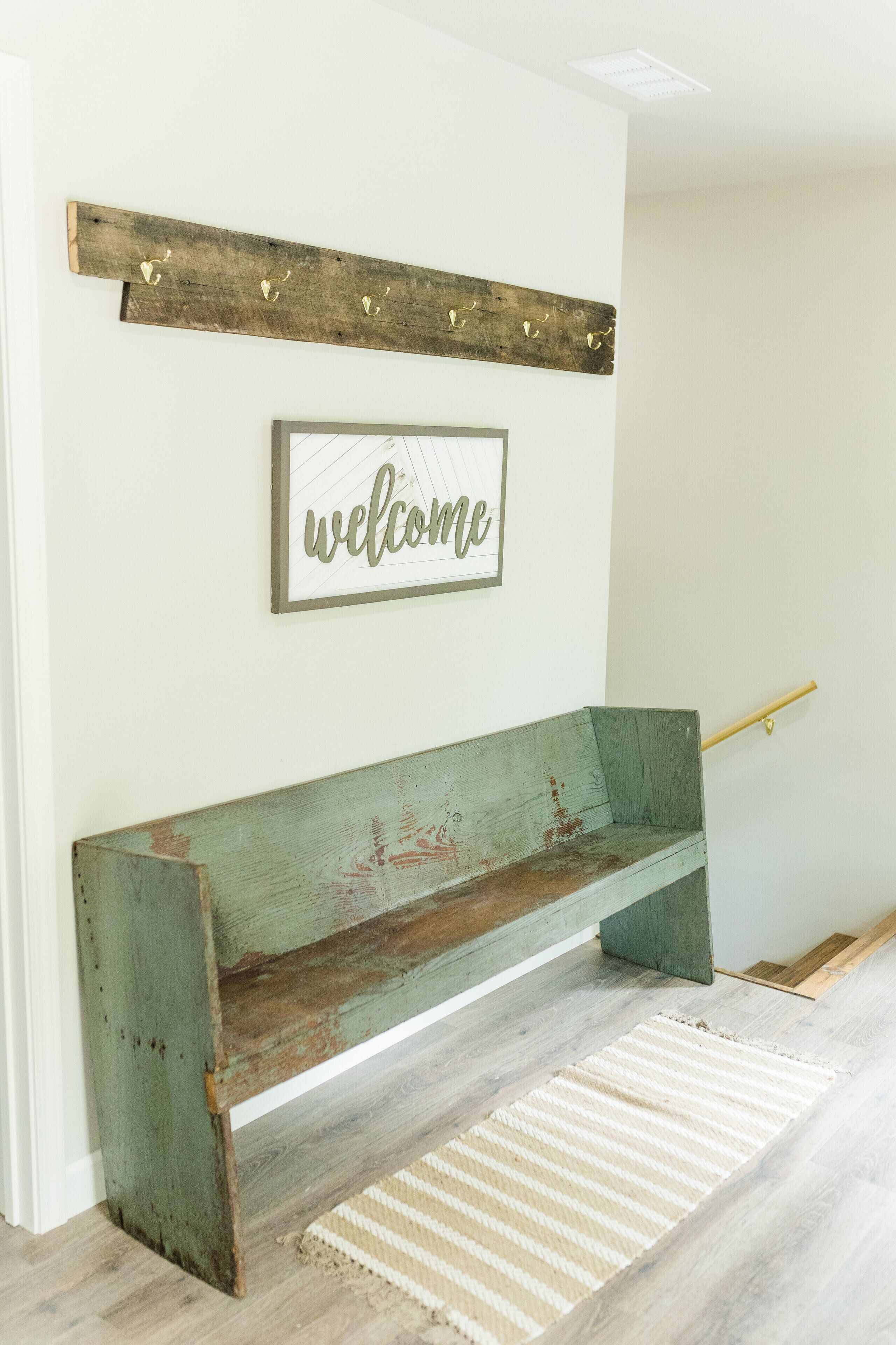 A rustic bench with a 'welcome' sign above it, a wooden beam, and a striped rug on a light-colored floor.