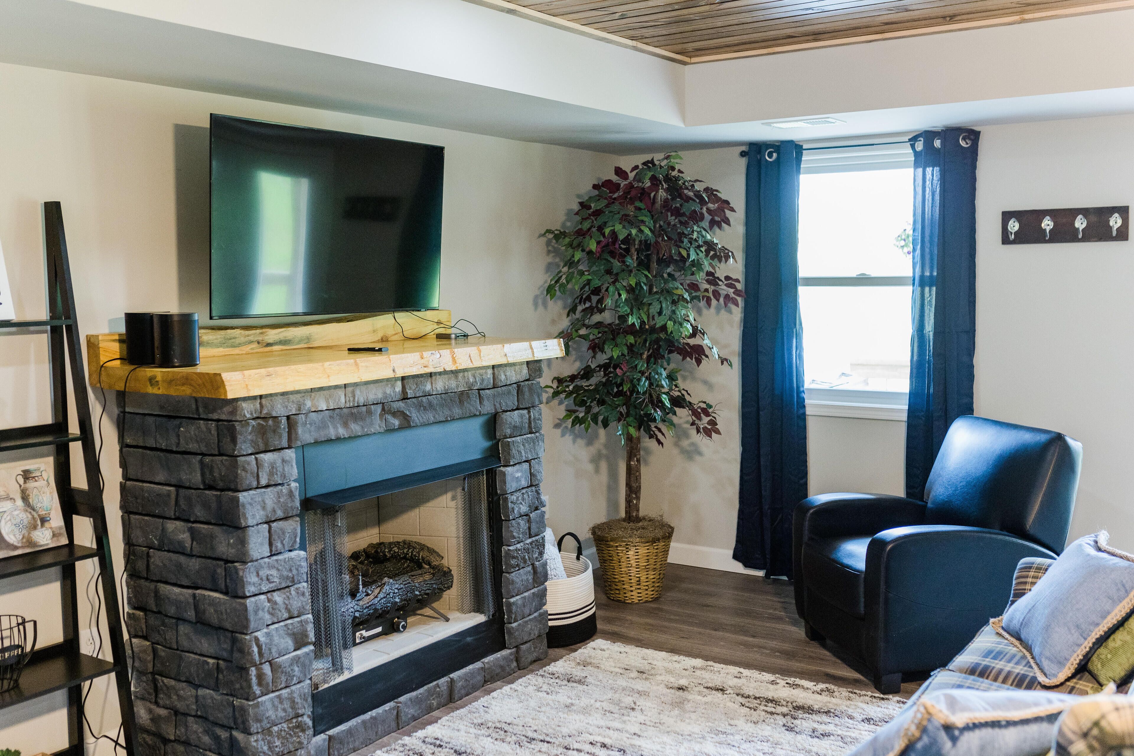 A living room with a stone fireplace, a mounted flat-screen TV, a large potted plant, a black leather chair, and a wooden floor.