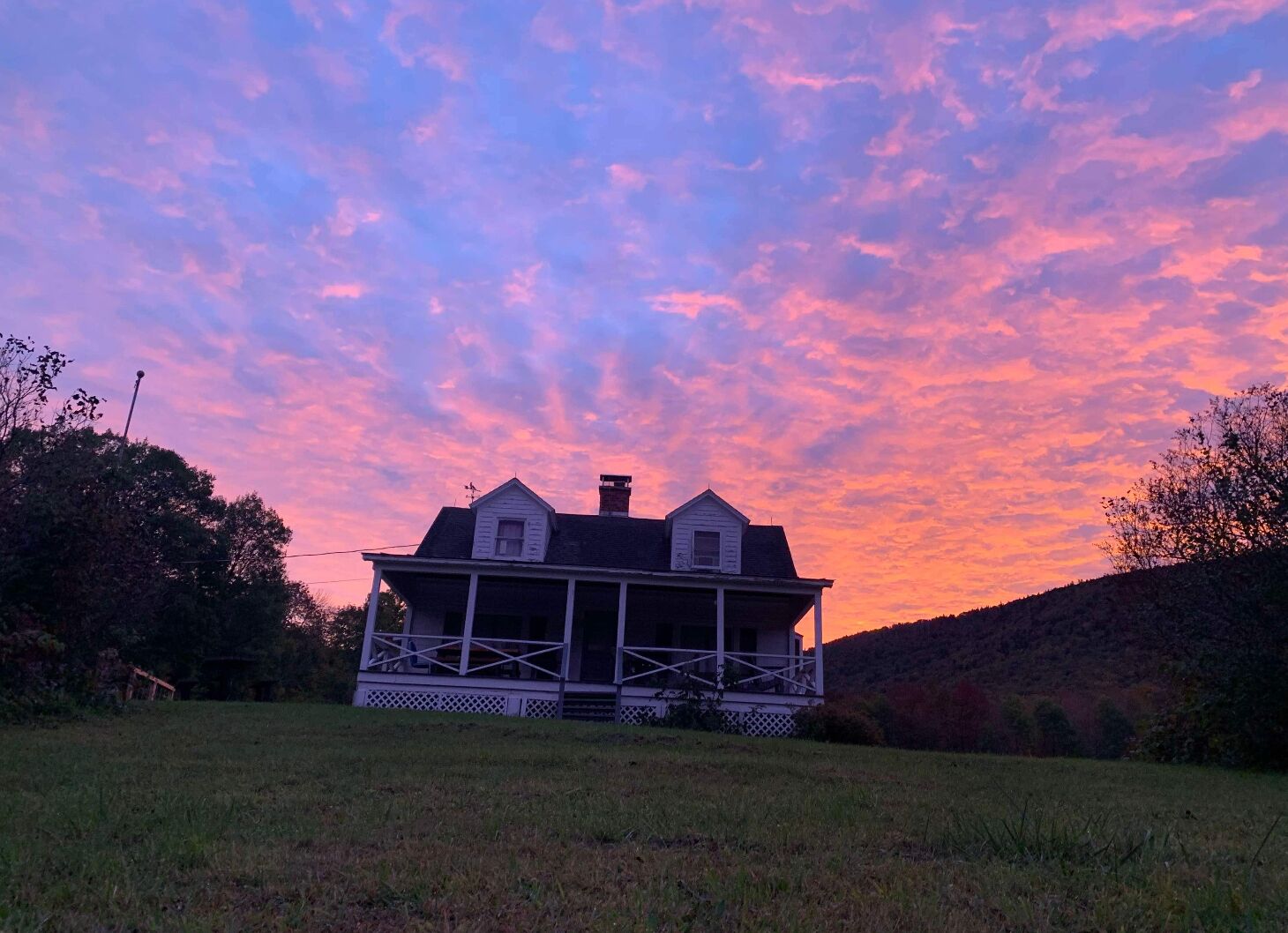 Una casa de dos pisos con un porche, rodeada por un campo verde y árboles, bajo un cielo con tonos de atardecer.