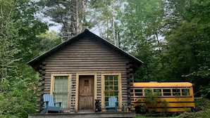 Eine rustikale Holzhütte mit Veranda, zwei Adirondack-Stühlen und einem in der Nähe geparkten Schulbus in Gelb.