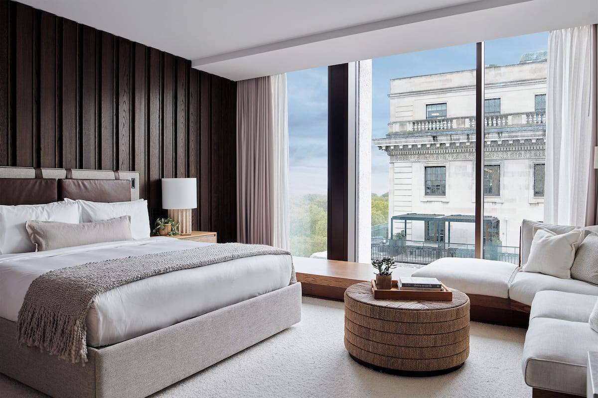 A modern bedroom with a large bed, a round ottoman, and a view of a cityscape through large windows.