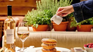 A table with a bottle of wine, a glass, a stack of donuts, and a hand pouring syrup. - 1 Hotel Mayfair (London)