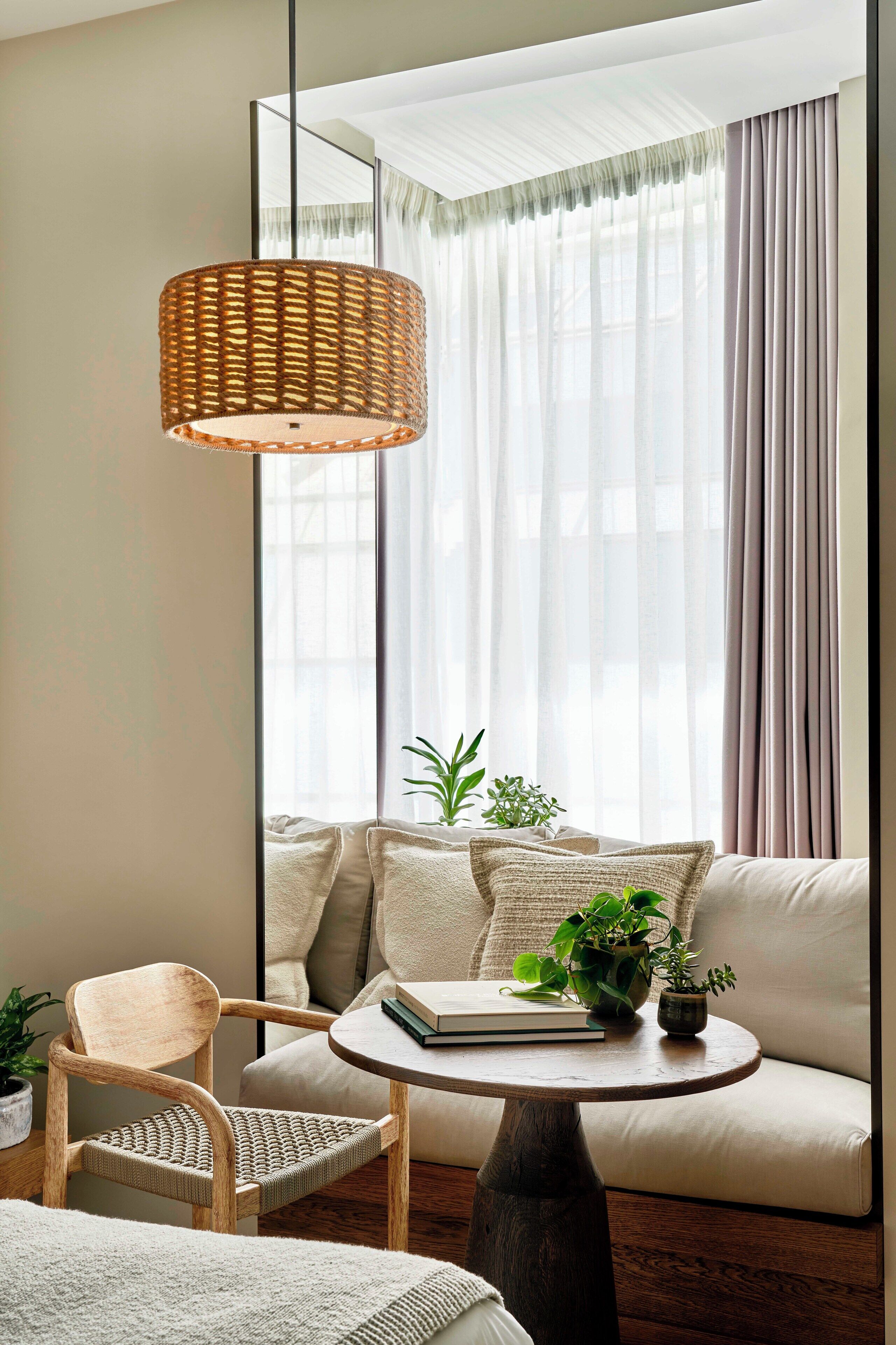 A modern living room with a large mirror, a wicker lamp, a sofa, a chair, and two potted plants.