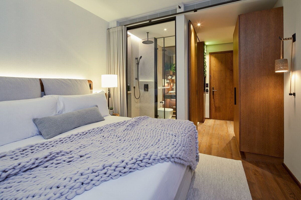 A modern hotel room with a large bed, a glass shower enclosure, and a wooden wardrobe.