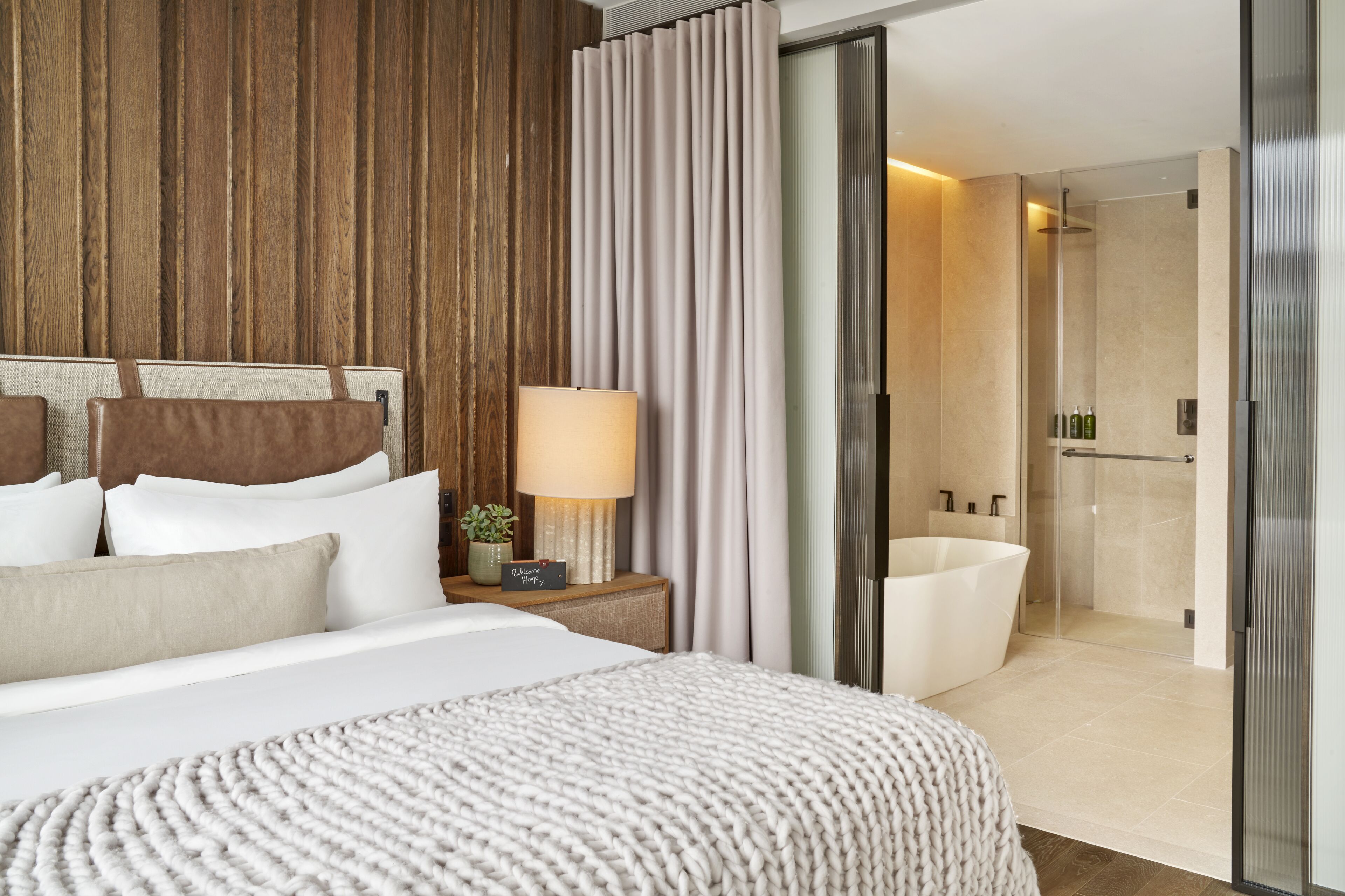 Une chambre d’hôtel moderne avec un grand lit, une table de chevet avec une lampe, une plante en pot et une vue sur une salle de bain équipée d’une baignoire et d’une douche.