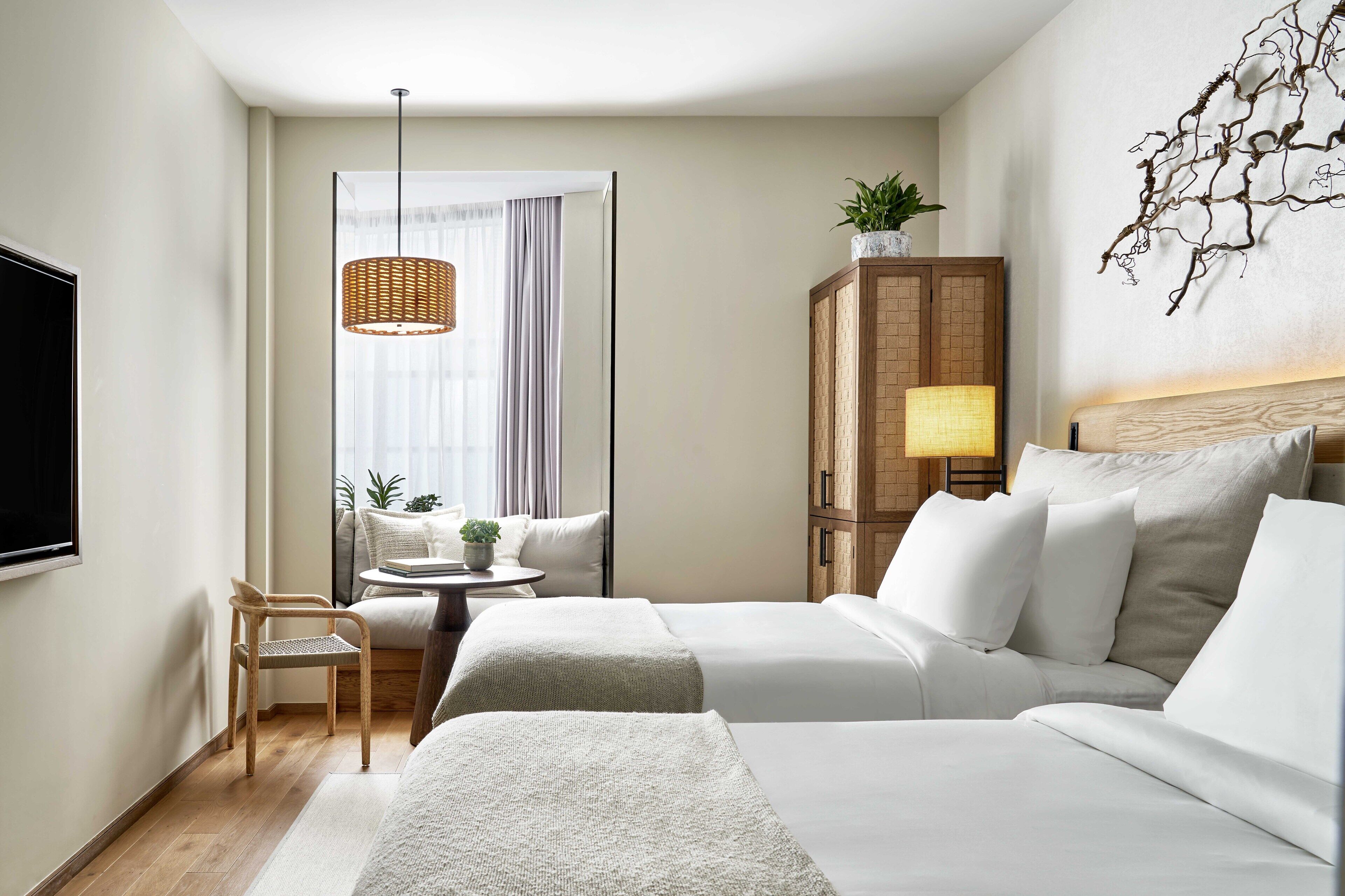 Een hotelkamer met twee bedden, een houten kast, een kleine tafel met een plant, een hanglamp van riet en een groot raam met gordijnen.