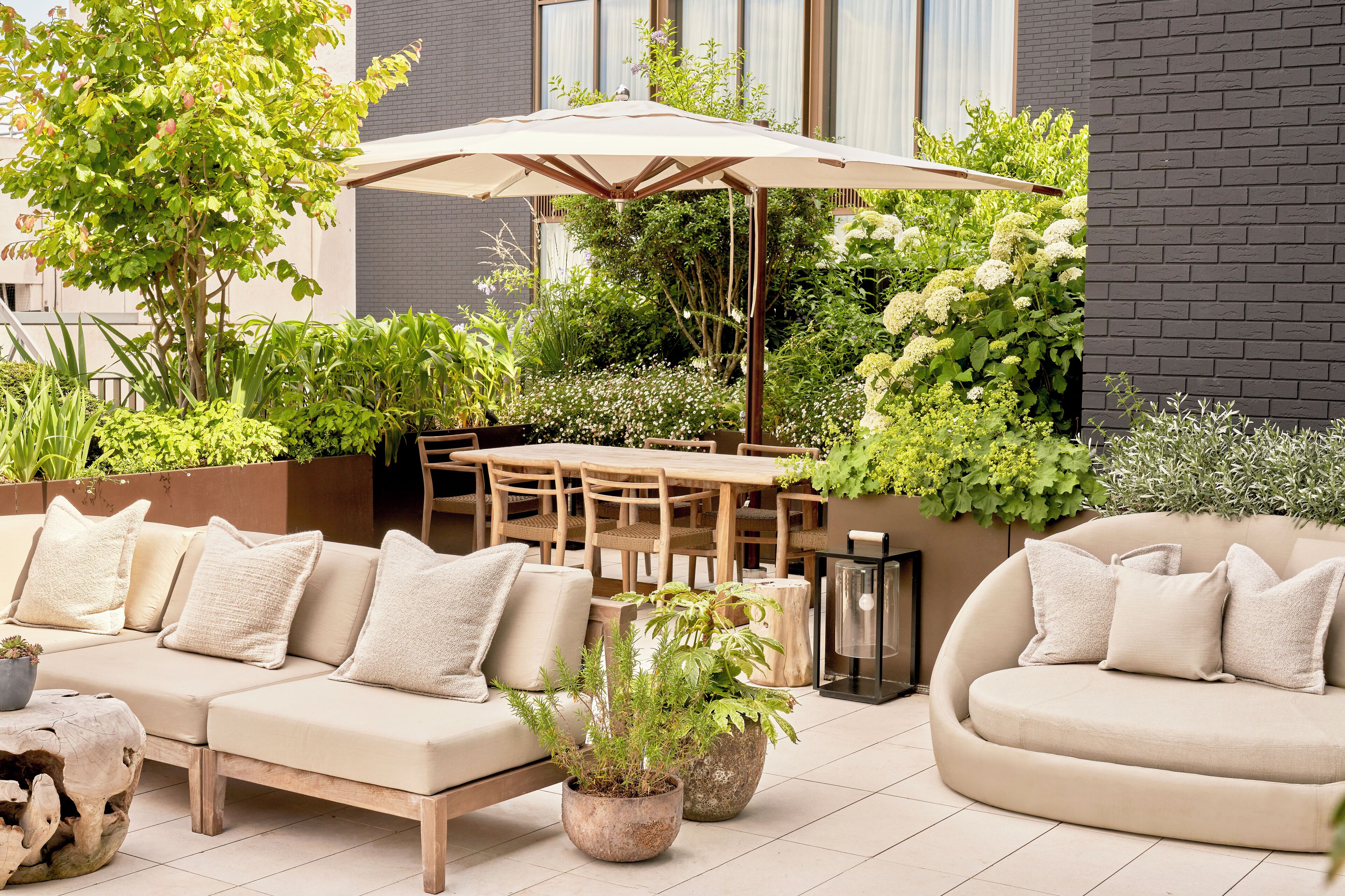 An outdoor patio area with a sofa, dining table, and chairs, surrounded by potted plants and a large umbrella.
