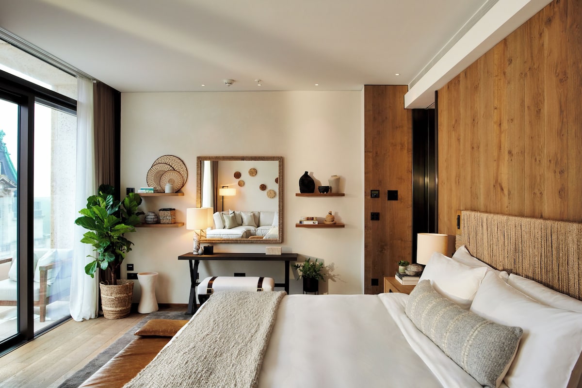 A modern hotel room with a large bed, a desk with a computer, a potted plant, and a view of the city.