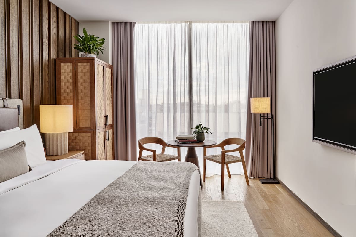 A hotel room with a bed, a desk with a lamp, a television, and a view of the city through sheer curtains.