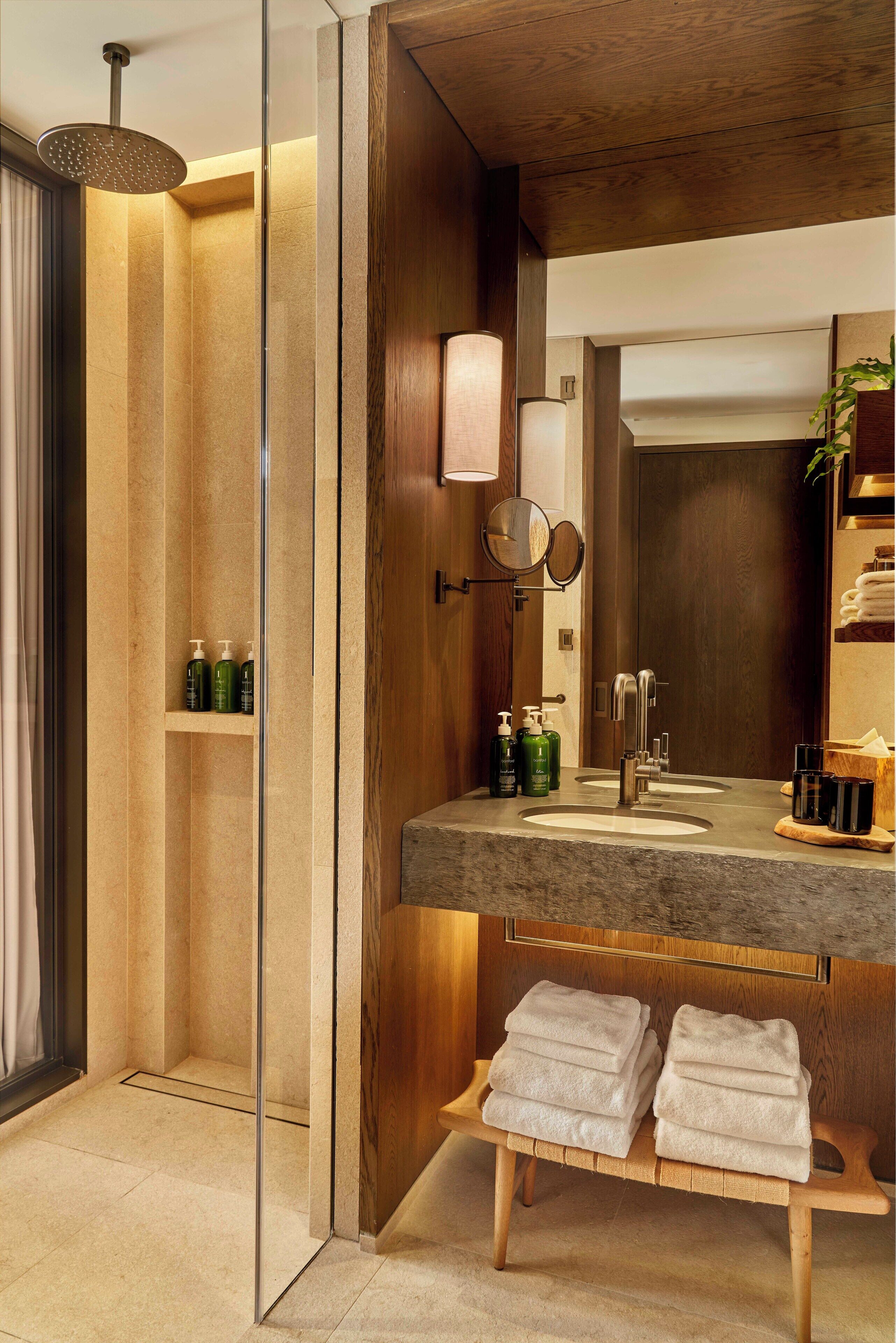 A modern bathroom with a glass-enclosed shower, a wooden vanity with a mirror, and a bench with towels.