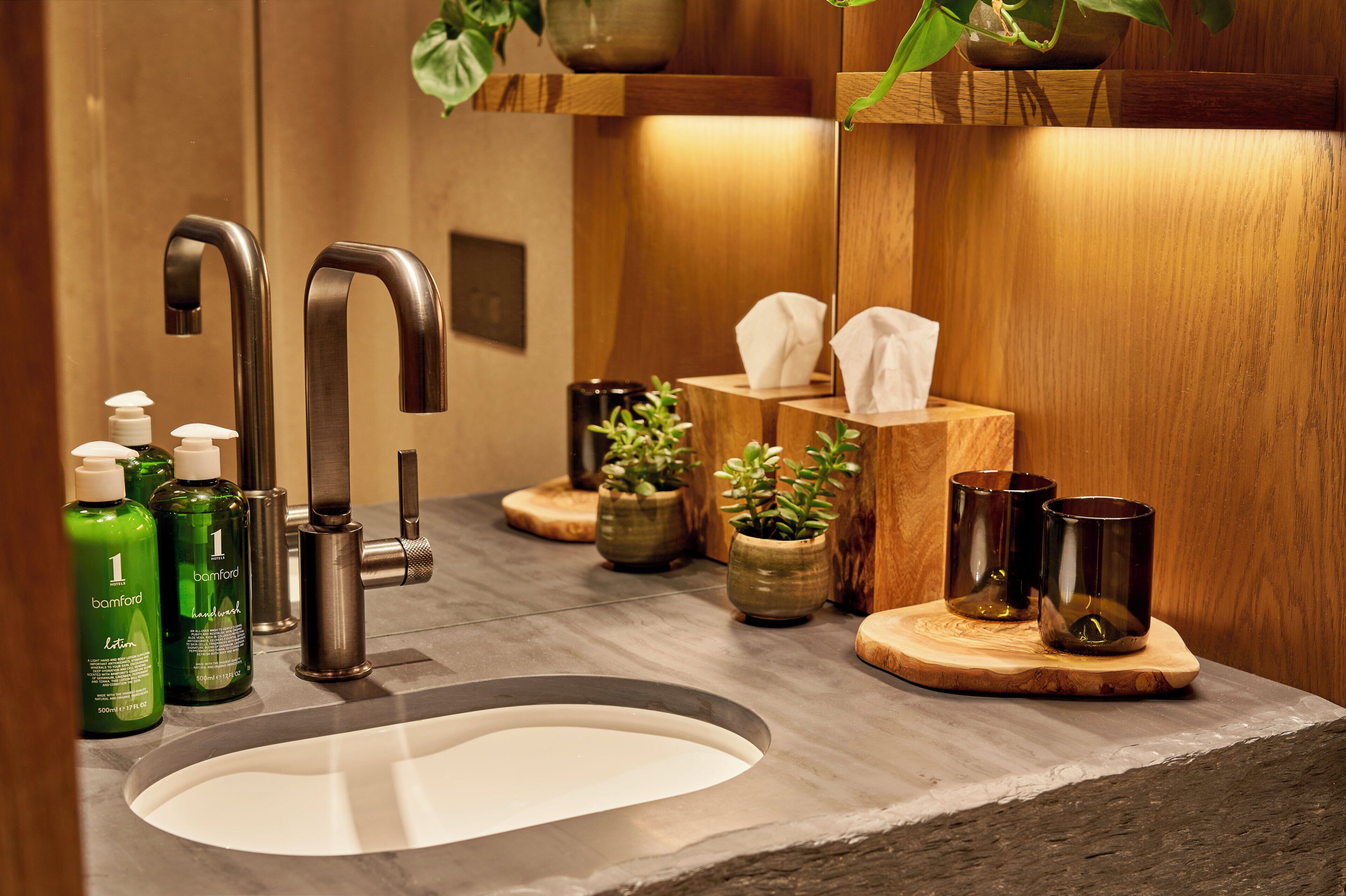 A bathroom with a modern sink, two faucets, a mirror, a wooden vanity, a potted plant, and toiletries.