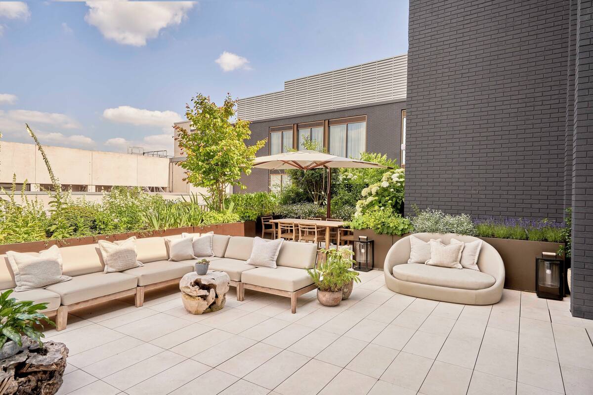 A rooftop patio with a long beige sofa, a round beige sofa, and a dining table with chairs. There is a large umbrella, potted plants, and a wooden table with a vase.