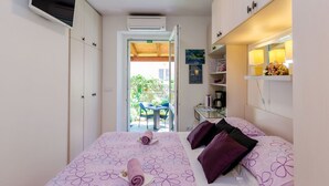 A hotel room with a bed, a TV mounted on the wall, an air conditioning unit, and a small table with a chair outside the door. - Apartments and Room Frane - Double Room (Dubrovnik)