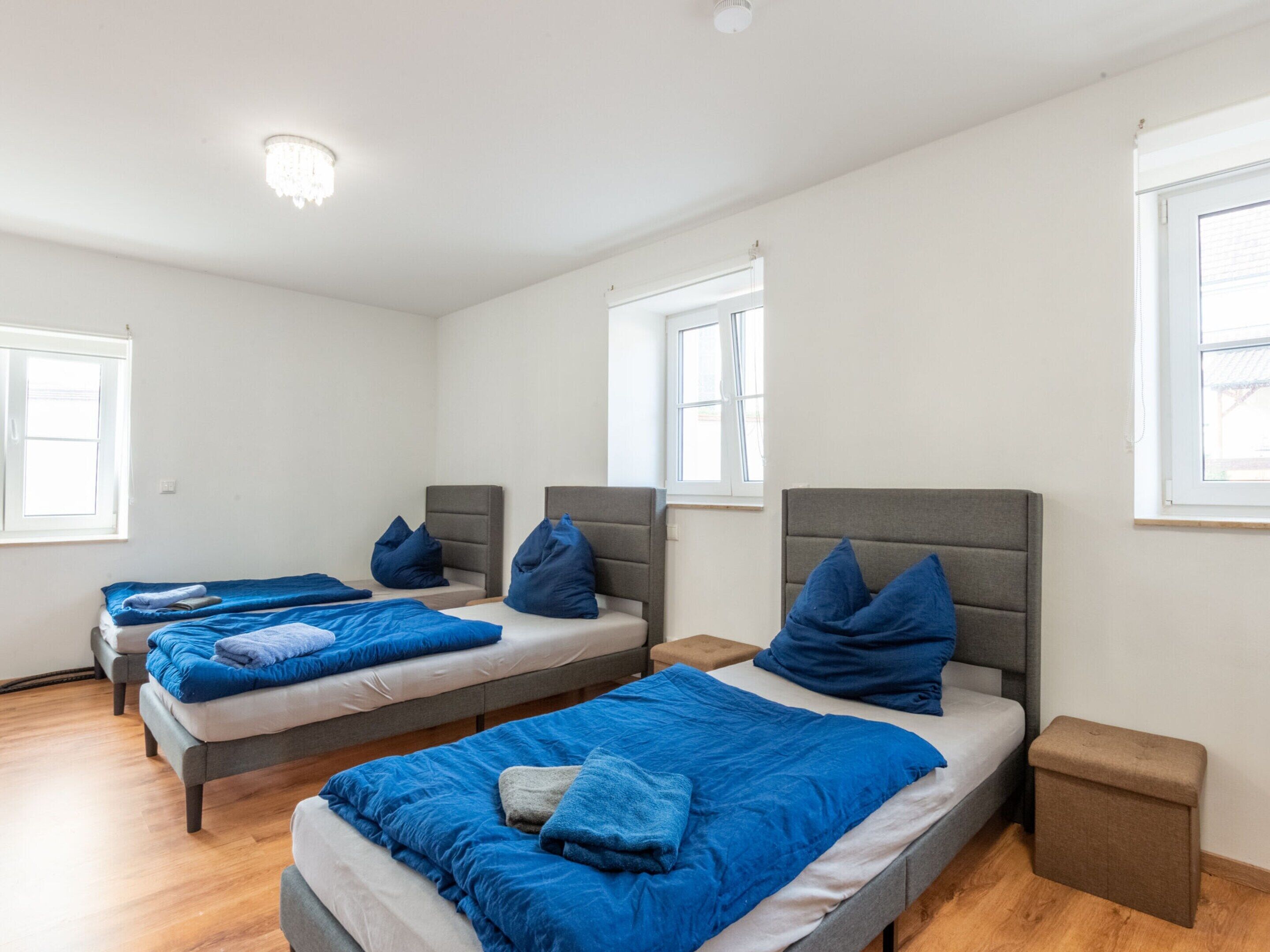 Habitación de hotel con tres camas individuales, cada una con cabecero gris y ropa de cama azul. Hay ventanas a dos lados de la habitación y un pequeño banco de madera junto a la cama derecha.