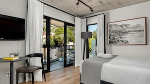 A modern hotel room with a bed, a desk with a chair, a TV, and a balcony with a view of the city and mountains. - Charming Apartment with Mountain Views (Cape Town)