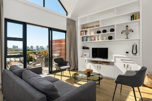 Una sala de estar moderna con un ventanal grande, sofás grises, una mesa de centro de vidrio y un mueble blanco de entretenimiento con libros y objetos decorativos.