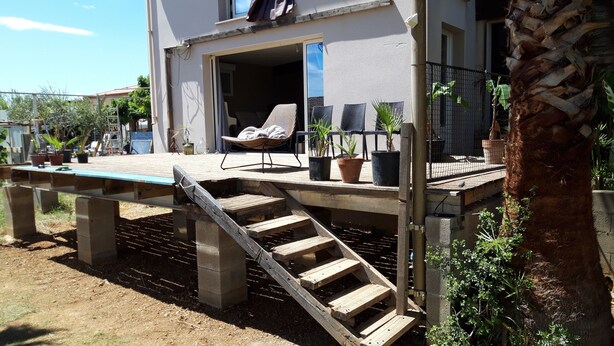 Une terrasse en bois avec une chaise et des plantes en pot, un escalier menant à un balcon et un palmier.