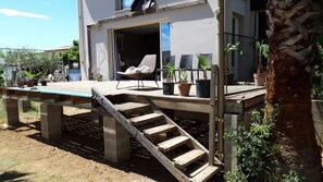 Eine Holzterrasse mit Stuhl und Topfpflanzen, eine Treppe, die zu einem Balkon führt, und eine Palme.