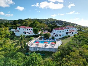 A hillside property with multiple white buildings, a swimming pool with sun loungers, and lush greenery.