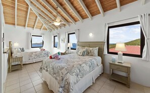 Une chambre à coucher avec un grand lit, deux tables de chevet, un ventilateur de plafond et une fenêtre donnant sur un beau paysage.