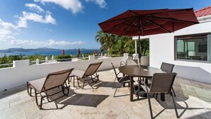 An outdoor patio with a round table and four chairs, a large red umbrella, and a view of the sea and mountains. - 2 bedroom, 2.5 bath with pool access & ocean view (Cruz Bay)