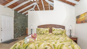 A bedroom with a wooden ceiling, a bed with a tropical leaf pattern bedspread, two bedside lamps, and a stone wall.