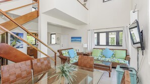 A spacious living area with a high ceiling, a wooden staircase, wicker furniture, a glass coffee table, and a television mounted on the wall.