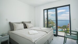 Een slaapkamer met een bed, nachtkastje, stoel en uitzicht op zee door een groot raam.