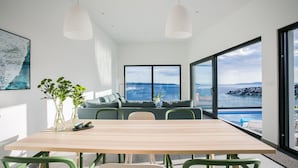 Een moderne eetruimte met een houten tafel en groene stoelen, een bank met kussens en grote ramen met uitzicht op zee.