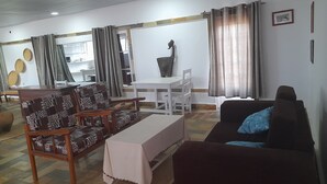 A living room with a sofa, a dining table, and chairs. There is a whiteboard and a window with curtains.
