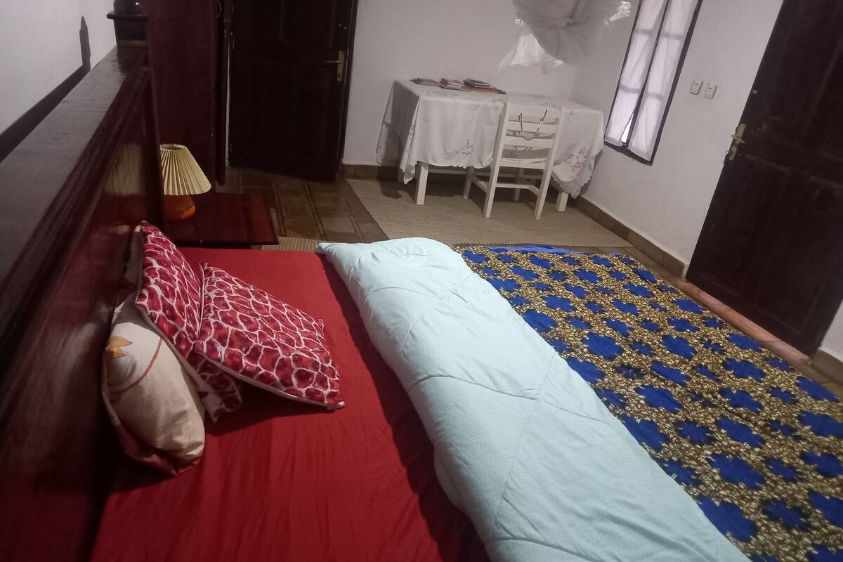 A bedroom with a red bedspread, a light blue blanket, and a patterned rug. There is a small table with a white tablecloth and a chair.