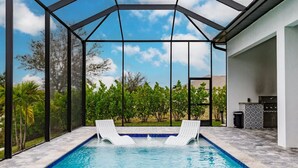 A swimming pool with two lounge chairs, surrounded by a glass enclosure with a view of trees and a clear sky.