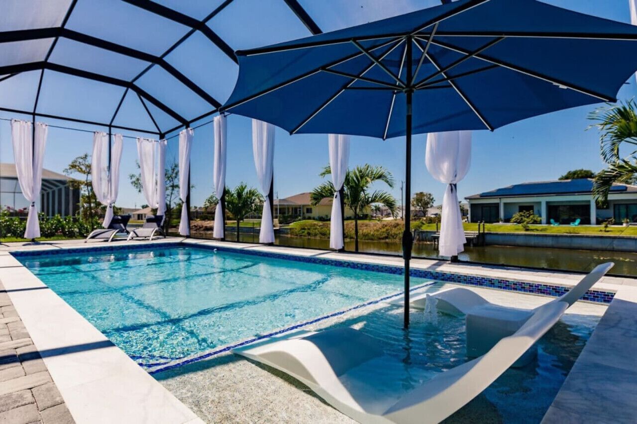Una piscina con cerramiento de vidrio, cortinas blancas y una sombrilla azul.