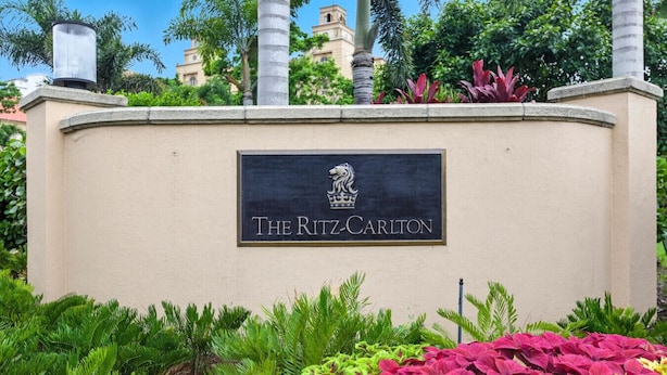 A sign for The Ritz-Carlton with a logo, surrounded by lush greenery and palm trees.
