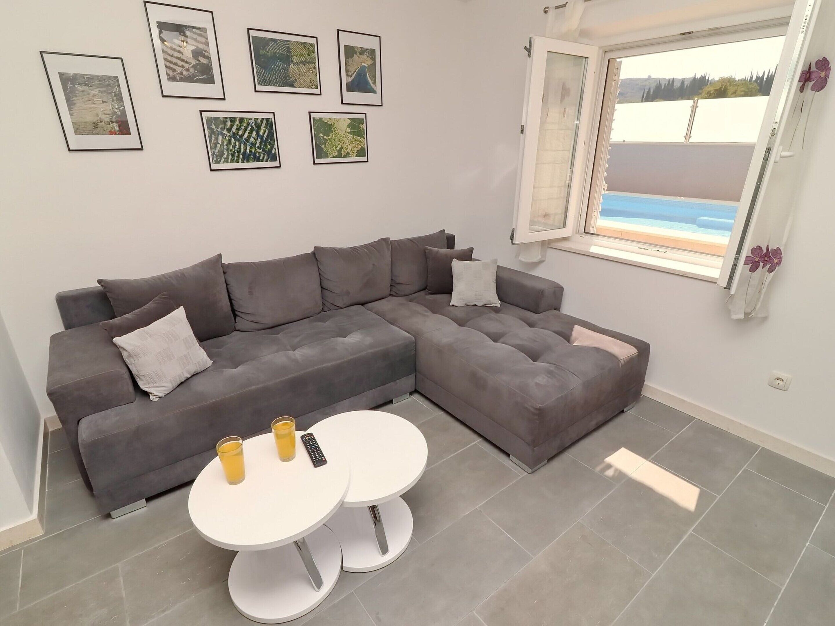 A modern living room with a grey sectional sofa, two white round tables, and a large window with a view of the sea.