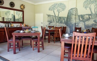 A dining area with a table set for four, surrounded by wooden chairs. A painting of trees and a brick wall decorates the wall behind the table.