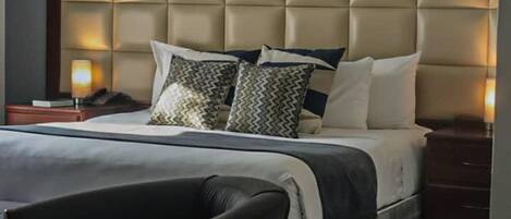 Un dormitorio moderno con una cabecera capitonada, una cama grande con muchas almohadas, una mesita de noche con una lámpara y una silla.