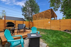 A backyard with a wooden deck, a grill, a table with chairs, and a fire pit.
