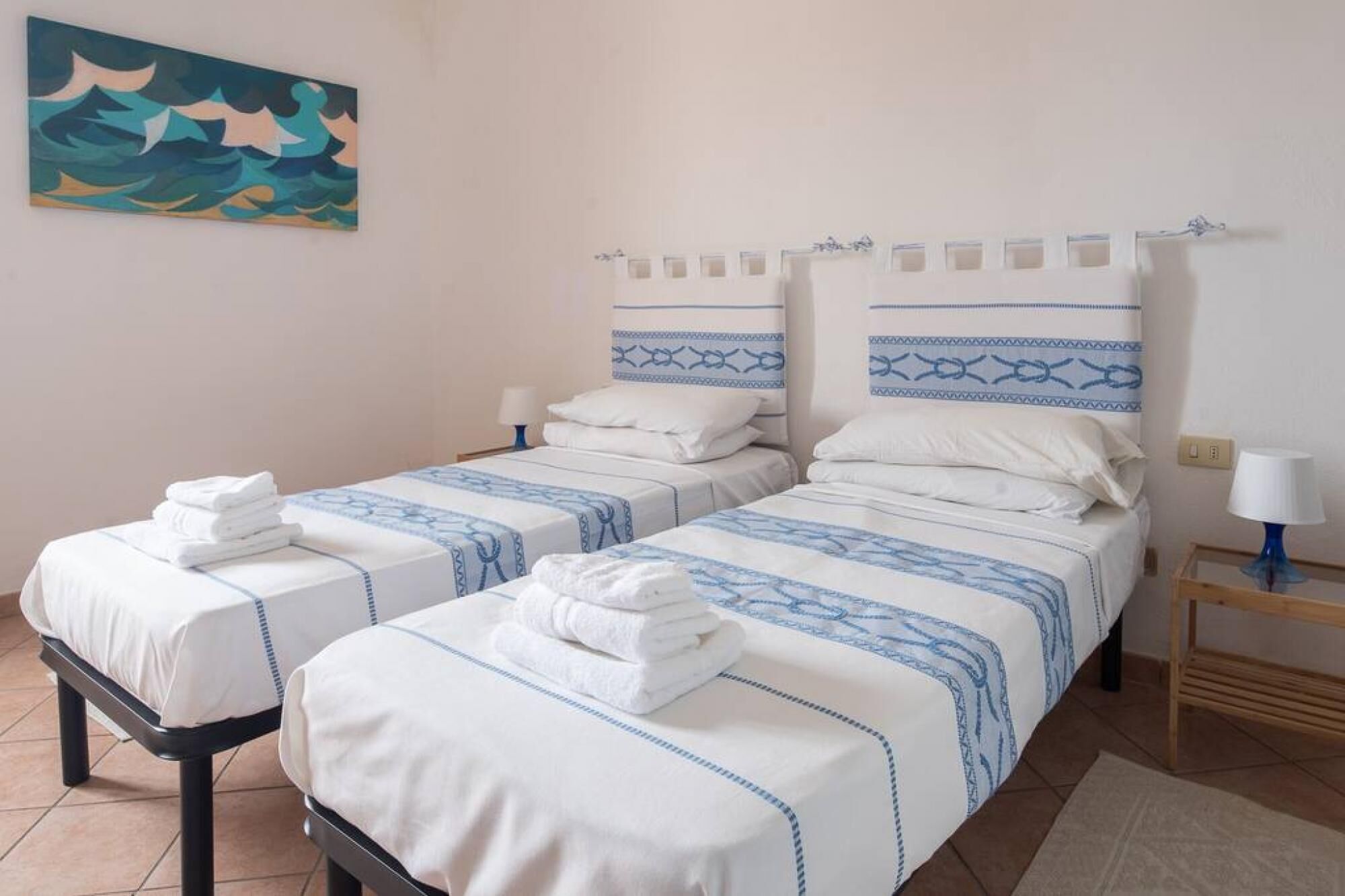 Ein Hotelzimmer mit zwei Betten, jeweils mit weißer Bettwäsche und blauen Mustern, einem Bild an der Wand und einem kleinen Tisch mit einer Lampe.
