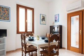 A dining area with a white table, wooden chairs, and a black cabinet. There are framed pictures on the walls and a TV on the left.